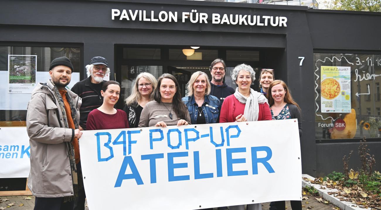 10 Personen stehen vor dem Pavillon. Sie lächeln und halten ein Banner hoch auf dem steht B4f Pop-Up-Atelier