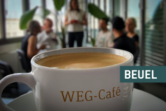 Kaffeetasse mit der Aufschrift "WEG-Café", im Hintergrund Menschen im Gespräch