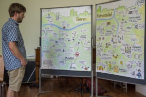 Ein junger Mann schaut sich 2 Pinwände an auf denen Illustrationen von einem Klimaneutralen Bonn hängen
